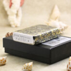 Decorative metal lighter with an intricate swirling pattern resting on a black box, with seashells in the background.
