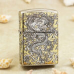 Gold-toned metal lighter with intricate black dragon design across front and sides, standing on a sandy surface with seashells nearby.