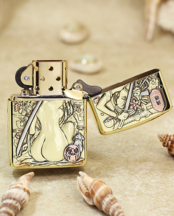 Gold Zippo lighter with intricate Japanese-style engravings and an open lid on a sandy surface with seashells nearby.