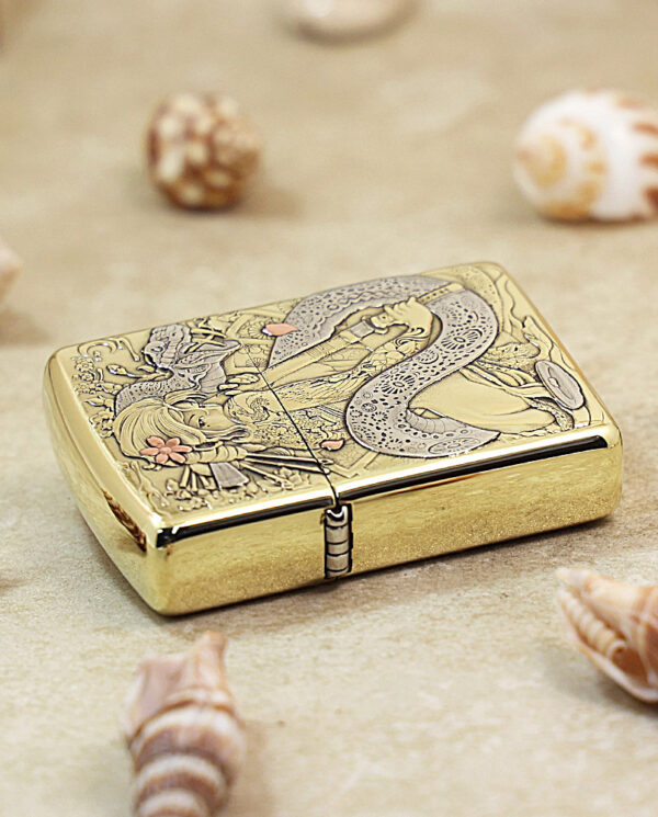 Gold metal lighter with an ornate dragon engraving, resting on a sandy surface among seashells.