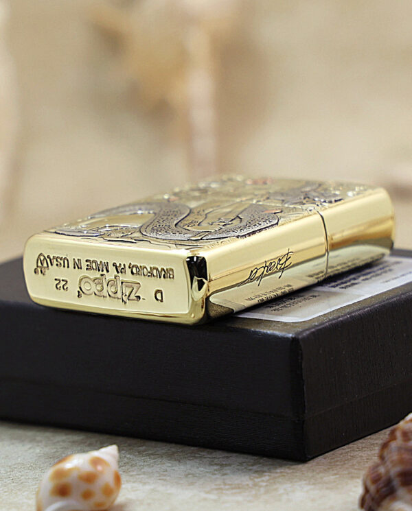 Gold engraved Zippo lighter resting on a black box on a sandy surface with small seashells nearby