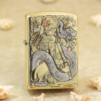 Gold Zippo-style lighter with an intricate engraving of a woman and a coiling dragon among flowers, set on a sandy background with seashells nearby.