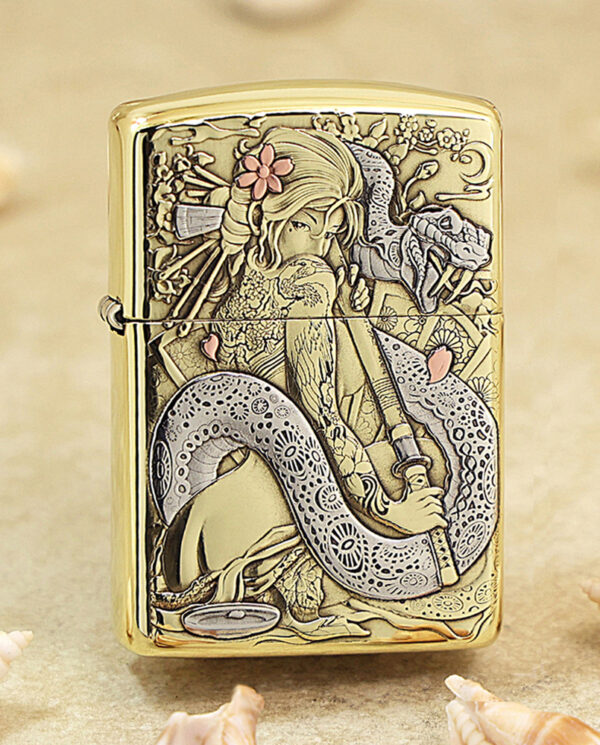 Gold Zippo-style lighter with an intricate engraving of a woman and a coiling dragon among flowers, set on a sandy background with seashells nearby.