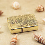Gold engraved lighter with a dragon design on a sandy surface, surrounded by seashells.