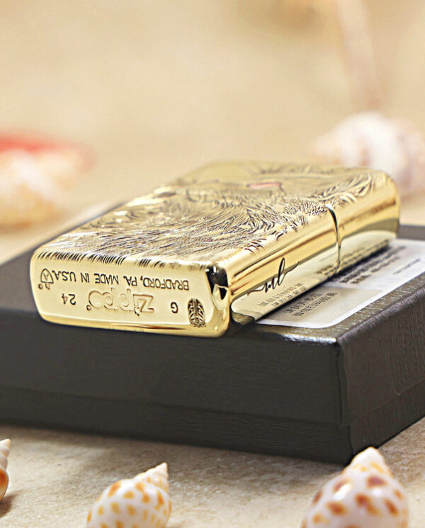 KZ152-05 Gold engraved Zippo lighter resting on a black box, with seashells scattered around in a soft-focus setting.