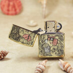 Gold Zippo-style lighter with ornate koi fish and floral engravings, lid open on a sandy surface with shells nearby