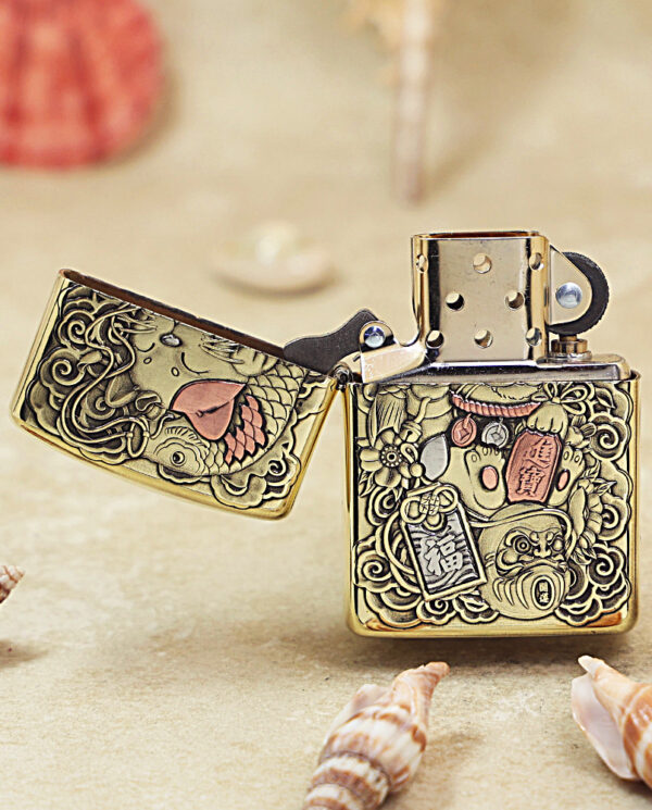Gold Zippo-style lighter with ornate koi fish and floral engravings, lid open on a sandy surface with shells nearby