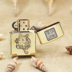 Gold Zippo lighter with a cute cat and flowers engraving, lid open, surrounded by seashells