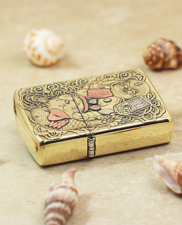 Gold decorative lighter with ornate engravings and a dragon motif, placed on sand with seashells nearby.