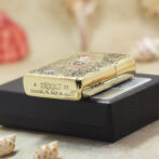 Gold Zippo lighter with engraved pattern resting on a black box, shells scattered nearby, soft beach setting.