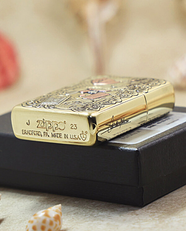 Gold Zippo lighter with engraved pattern resting on a black box, shells scattered nearby, soft beach setting.