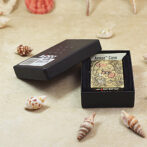 Ornate gold Zippo lighter in a black box with Armor Case label on a sandy surface among seashells