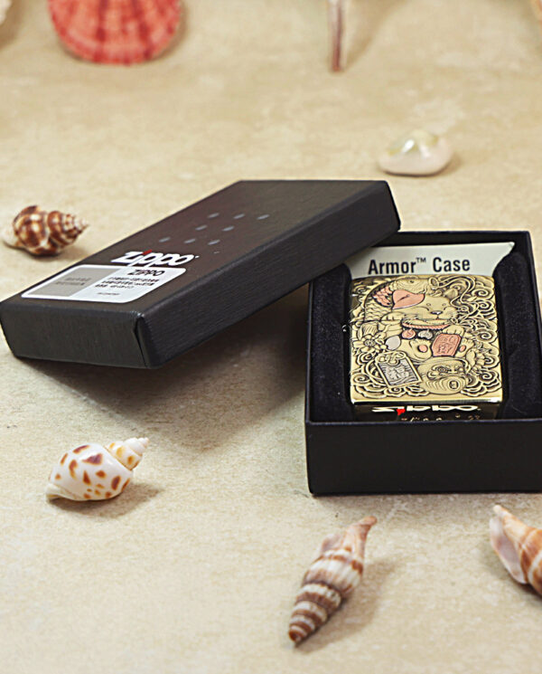 Ornate gold Zippo lighter in a black box with Armor Case label on a sandy surface among seashells