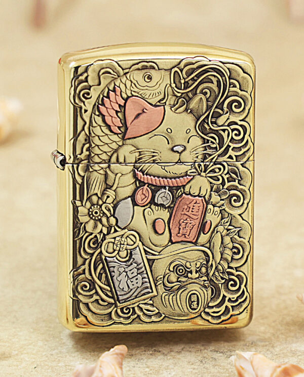 Gold Zippo lighter with an embossed Maneki-neko (lucky cat) design and hanging charms, ornate and decorative.
