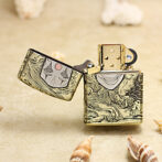 Gold Zippo lighter with ornate engraving and open lid, featuring a stylized face and waves on a sandy background.