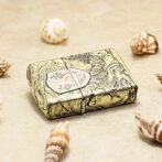Ornate gold-tone lighter with a detailed engraving of a face, placed on sand among seashells.