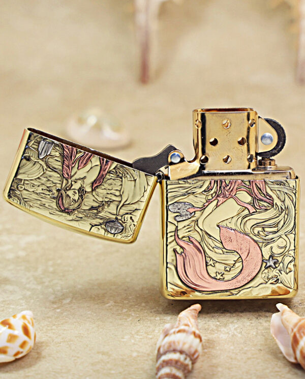 Gold Zippo-style lighter with mermaid artwork, lid open, on sandy beach with seashells nearby.