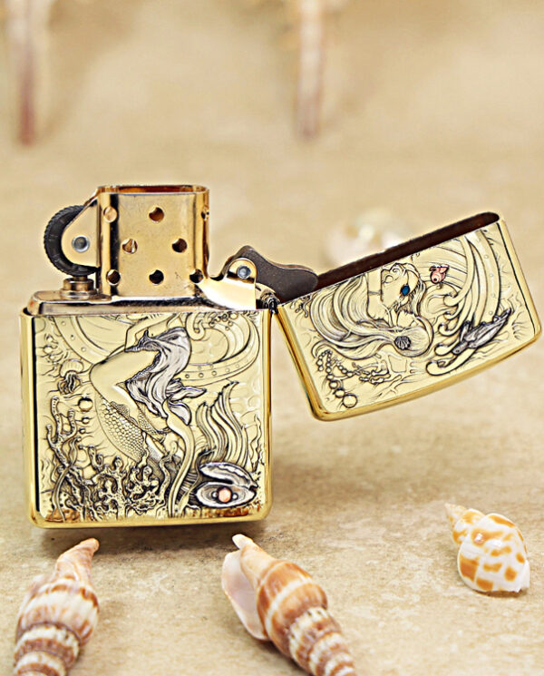 Gold engraved Zippo lighter with a mermaid sea motif, open lid, resting on sandy beach with seashells nearby.