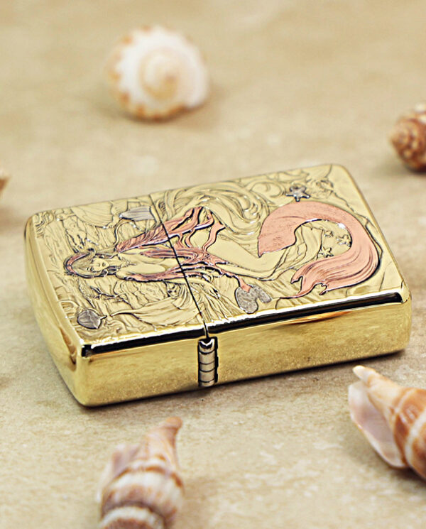 Gold engraved lighter on a sandy surface, featuring a mermaid among seaweed with seashells around it.