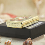 Gold Zippo lighter with engravings resting on a black box, surrounded by seashells on a sandy surface.