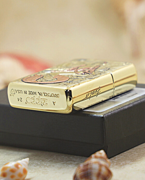 Gold Zippo lighter with engravings resting on a black box, surrounded by seashells on a sandy surface.