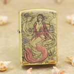 Gold-tone lighter with an engraved mermaid undersea scene, pink tail, and swirling waves surrounded by seashells and coral contrasts on a sandy background.