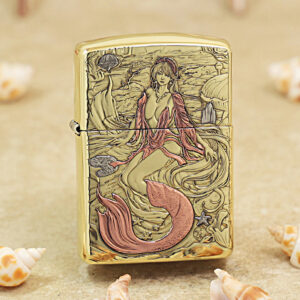 Gold-tone lighter with an engraved mermaid undersea scene, pink tail, and swirling waves surrounded by seashells and coral contrasts on a sandy background.