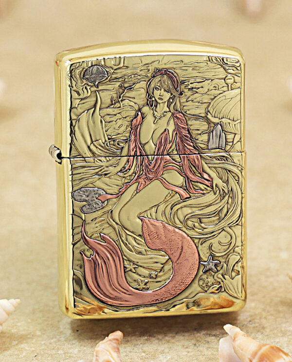 Gold-tone lighter with an engraved mermaid undersea scene, pink tail, and swirling waves surrounded by seashells and coral contrasts on a sandy background.