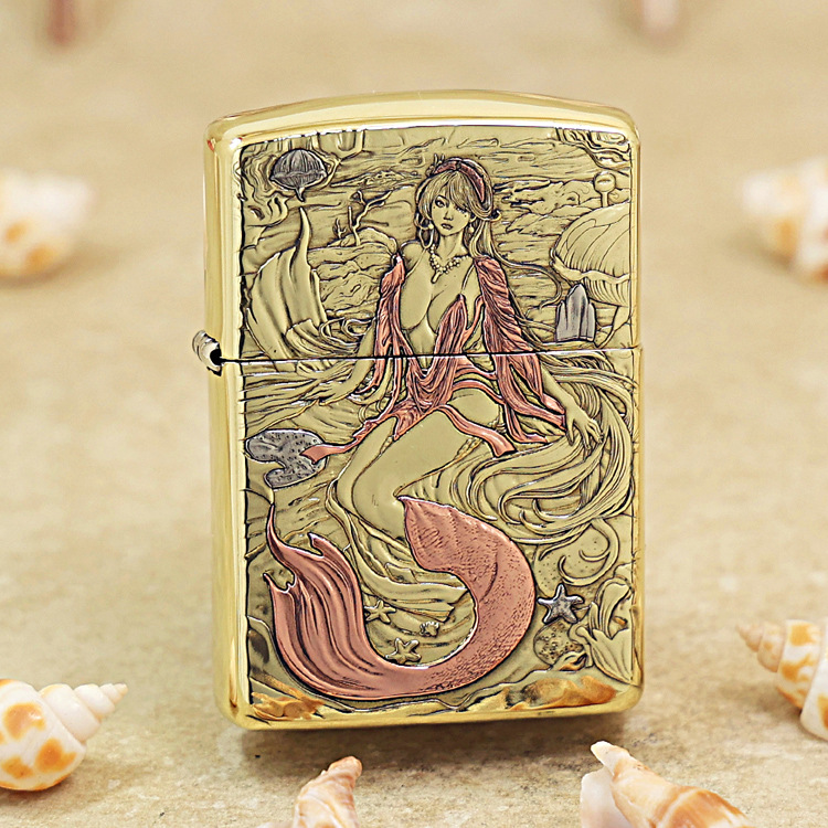 Gold-tone lighter with an engraved mermaid undersea scene, pink tail, and swirling waves surrounded by seashells and coral contrasts on a sandy background.
