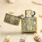 Brass Zippo-style lighter with an intricate elephant engraving, lid open, on a sandy surface with seashells nearby.