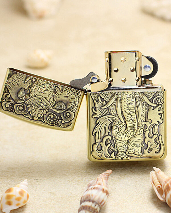 KZ158-02 Brass Zippo-style lighter with an intricate elephant engraving, lid open, on a sandy surface with seashells nearby.