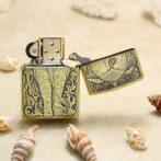 Brass Zippo lighter with intricate elephant engraving, open lid, on a sandy beach among seashells.
