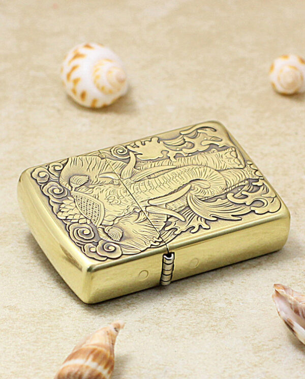 KZ158-04 Gold-toned metal lighter with an intricate dragon engraving, closed, resting on a beige surface with seashells nearby, decorative and ornate.