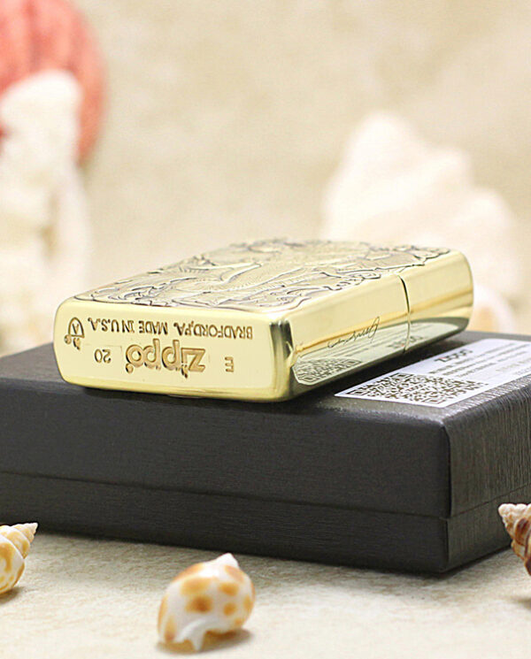 KZ158-05 Gold engraved Zippo lighter resting on a black box, surrounded by seashells on a light surface.