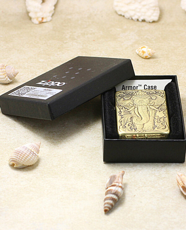 KZ158-06 Gold-tone Zippo Armor Case lighter in an open black presentation box on a sandy surface with seashells nearby, showcasing the engraved design on the lighter's front.