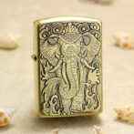 Brass Zippo-style lighter with a detailed embossed elephant deity design, placed on a sandy surface with seashells nearby.