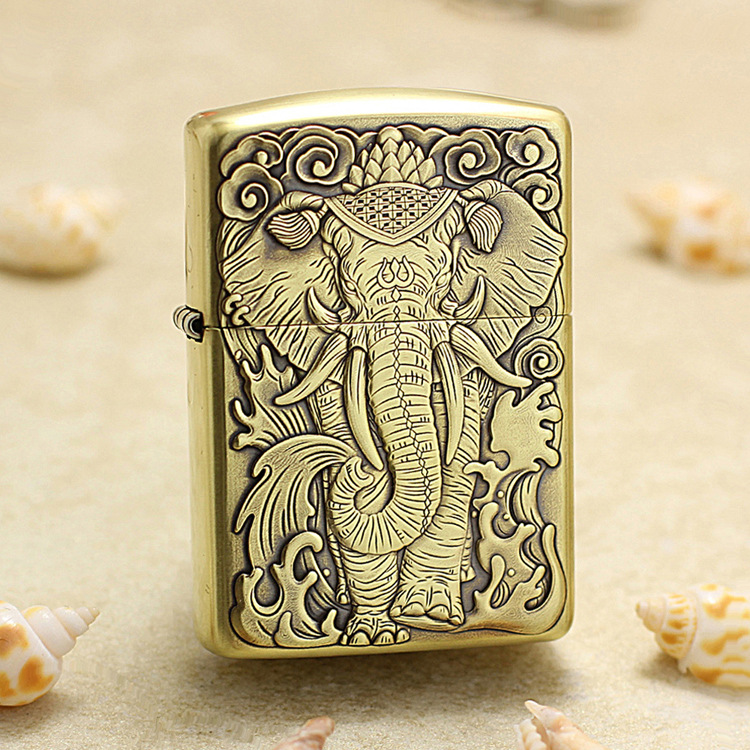 Brass Zippo-style lighter with a detailed embossed elephant deity design, placed on a sandy surface with seashells nearby.