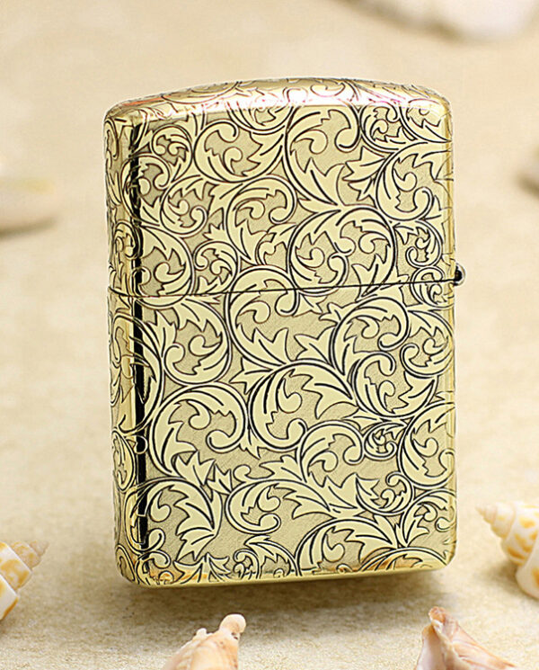 KZ159-01 Gold-toned Zippo-style lighter with intricate swirling leaf pattern on a sandy beach with seashells nearby