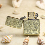 Gold Zippo lighter with an intricate floral pattern, open on a sandy beach with seashells nearby.