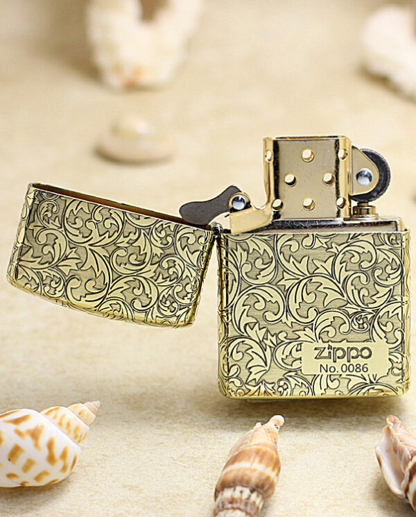 KZ159-02 Gold Zippo lighter with an intricate floral pattern, open on a sandy beach with seashells nearby.