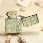 Gold patterned metal lighter with the lid open, resting on a sandy beach among seashells.