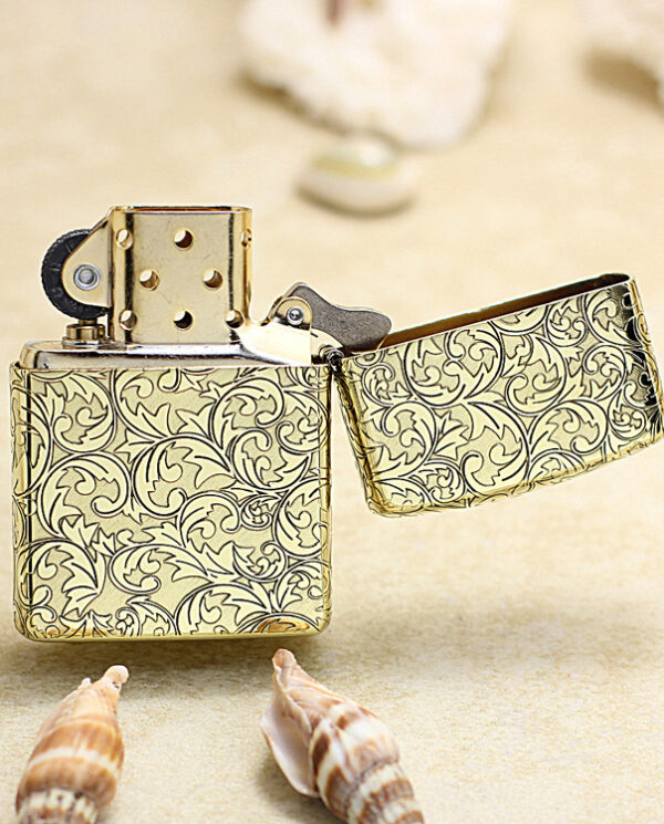 KZ159-03 Gold patterned metal lighter with the lid open, resting on a sandy beach among seashells.