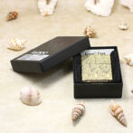 Gold patterned Zippo lighter in a black gift box on a sandy surface with seashells scattered around.