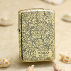 Zippo lighter in gold with ornate scrolling floral pattern, standing on sand with seashells nearby.