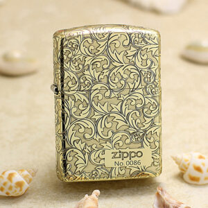 Zippo lighter in gold with ornate scrolling floral pattern, standing on sand with seashells nearby.