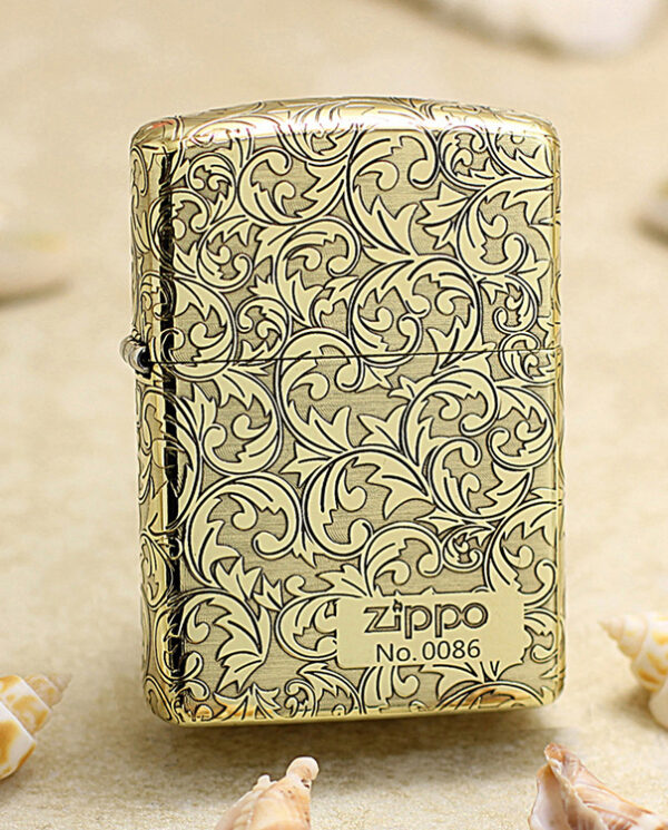 KZ159 Zippo lighter in gold with ornate scrolling floral pattern, standing on sand with seashells nearby.