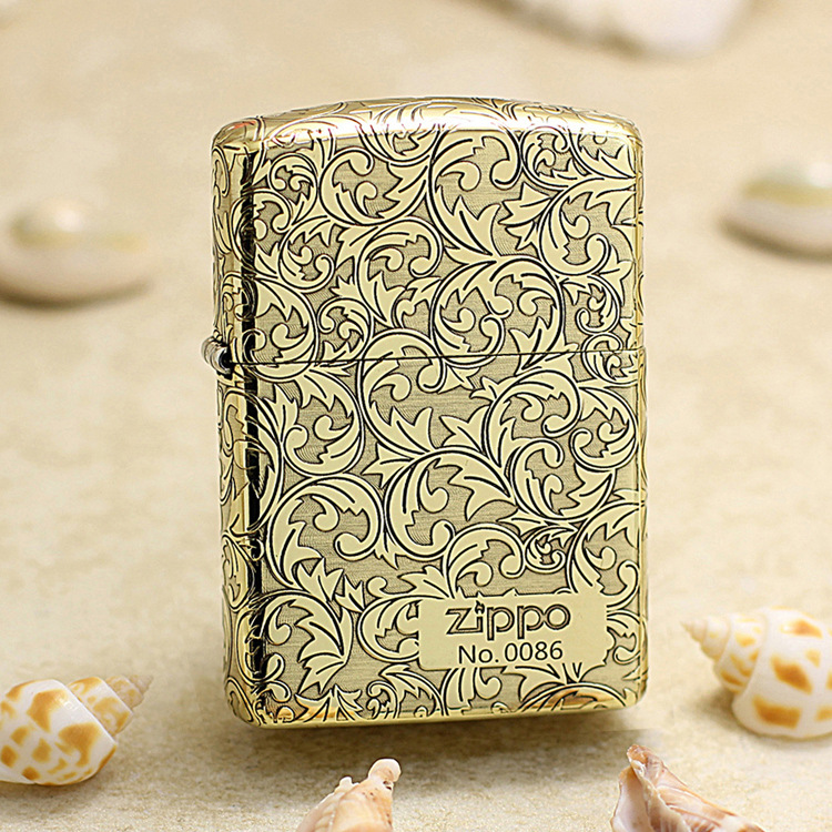 Zippo lighter in gold with ornate scrolling floral pattern, standing on sand with seashells nearby.