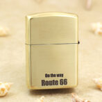 Brass Zippo-style lighter standing upright with 'On the way Route 66' printed on its front, on a sandy surface with tiny seashells nearby.