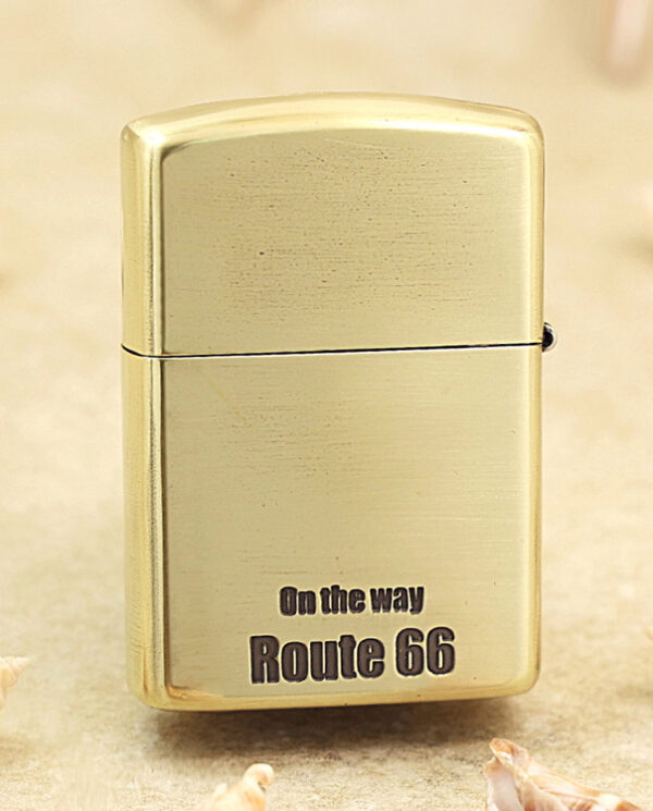 KZ160-01 Brass Zippo-style lighter standing upright with 'On the way Route 66' printed on its front, on a sandy surface with tiny seashells nearby.