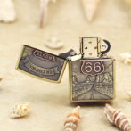 Bronze Zippo lighter with Route 66 shield and desert road engraving, lid open, on a sandy surface with seashells nearby.